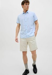 Cămașă cu mânecă scurtă de culoare albastru deschis, cu guler, asortată cu pantaloni scurți beji rulați și pantofi negri, completată de șosete gri.