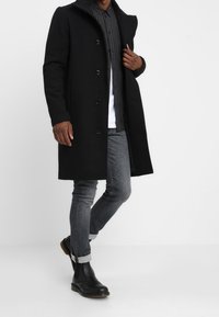 Uomo che indossa un soprabito nero, una camicia a quadri scuri, una maglietta bianca, jeans grigi arrotolati e stivali Chelsea neri, mentre posa contro uno sfondo chiaro e uniforme.
