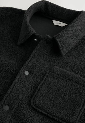 Svart texturerad fleecejacka med krage, tryckknappar och en fyrkantig bröstficka, som visar tygdetalj och varumärkesetikett på insidan.