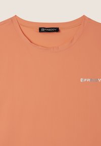 Camiseta de manga corta color durazno hecha de tela suave, con un escote redondo y un pequeño logo metálico en el lado izquierdo del pecho.