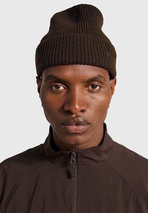 Mørkebrun ribstrikket hue med en tilspidset top, båret sammen med en matchende højhalset jakke med lynlås i et struktureret stof.