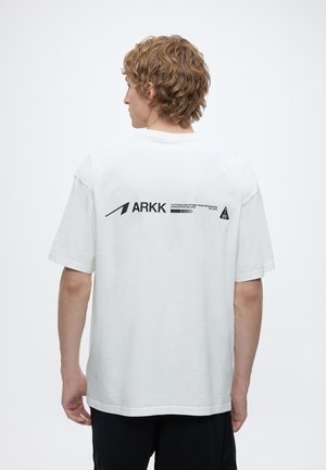 Personne aux cheveux courts et bouclés vue de dos portant un t-shirt blanc ARKK avec un texte noir et des graphismes sur le haut du dos.