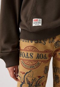 Felpa marrone con polsini a costine e etichetta Levi's; abbinata a pantaloni gialli con stampa e dettagli a tema giocattolo rossi e blu.
