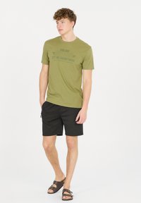 Olivgrön bomulls-t-shirt med svart tryckt text, kombinerad med svarta shorts och bruna sandaler; korta ärmar och avslappnad passform.