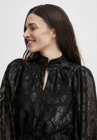 Bluză neagră cu un guler înalt, detaliu cu spărtură și un model texturat de leopard în negru metalic. Mânecile puff transparente adaugă volum.