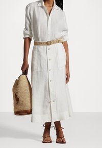 Vestido de lino blanco abotonado con una silueta de longitud media, ceñido con un cinturón tejido beige. También se ve un bolso tote de mimbre color tan.