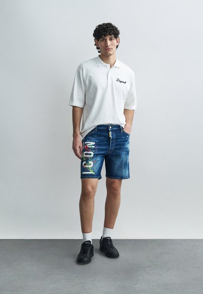 Polo blanc avec broderie noire, short en denim avec un motif graphique multicolore. Baskets noires et chaussettes blanches complètent le look.