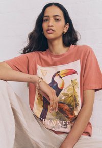 Woman with long dark hair wearing hoop earrings and a brick-red T-shirt featuring a colorful toucan and beige pants, seated against a white wall.