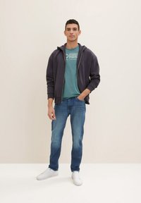 Mørk marineblå lynlås-hoodie over en teal grafisk t-shirt, parret med blå jeans og hvide sneakers; afslappet design med en løs pasform.
