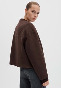 Jonge vrouw met slicked-back haar, gekleed in een losse donkerbruine wollen jas en zwarte jeans, kijkt over haar schouder.