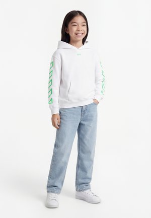 Smiling girl standing with one hand in pocket, wearing white hoodie with green sleeve designs, light blue jeans, and white sneakers.