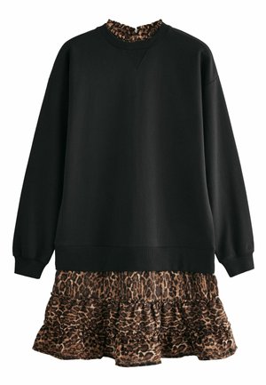 Persona vistiendo una blusa negra de manga larga y una falda de estampado de leopardo en capas, de pie con un brazo cruzado sobre la cintura frente a un fondo simple.