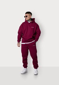 Hoodie bordeaux avec poche avant et capuche à cordon, associé à un pantalon de survêtement assorti. Accent blanc en dessous ; porté avec des baskets blanches.