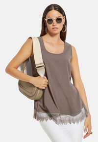 Braunfarbenes, ärmelloses Oberteil mit einem Rundhalsausschnitt, verziert mit einer Spitzenborte am Saum. Kombiniert mit einer beigen Umhängetasche und einer Sonnenbrille.