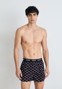 Un caleçon noir avec un motif répétitif de logo "HUGO" en rouge et blanc. Ceinture élastique avec détail du logo. Texture de tissu lisse.