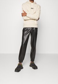 Kremowa bluza z kapturem z logo, w połączeniu z czarnymi joggerami z eko skóry z elastycznymi mankietami oraz czarnymi sneakersami z żółtymi akcentami.