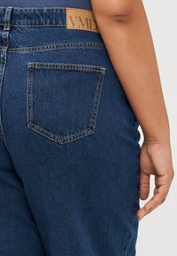 Pogled od blizu na zadnji žep temno modrih jeans hlač in stran človekove roke, ki počiva ob stegnu.