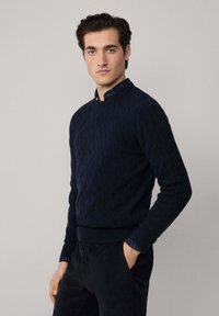 Maglione navy a trecce con scollatura rotonda, polsini e orlo a coste, indossato sopra una camicia a righe con colletto che presenta accenti in navy e bordeaux.