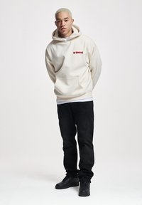 Felpa color crema con logo rosso, dotata di tasca frontale e cappuccio con coulisse, abbinata a jeans neri e sneaker nere.