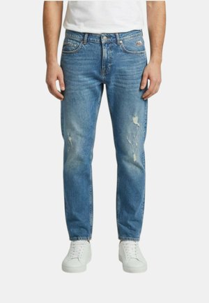 Uomo con jeans azzurri strappati e sneaker bianche, in piedi su uno sfondo bianco uniforme.