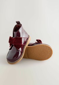 Glänzende burgunderfarbene Lacklederstiefel mit einem samtigen Schleifendetail, runder Zehenform und strukturiertem Gummisohle. Das Innere hat ein kariertes Muster.