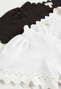 Deux hauts en coton, un noir et un blanc, avec des bords festonnés et des manches bouffantes, disposés à plat et montrant les étiquettes de marque et de taille.