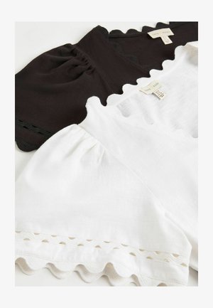 Dos camisetas de algodón, una negra y otra blanca, con bordes festoneados y mangas abullonadas, dispuestas en plano mostrando las etiquetas de la marca y las etiquetas de tamaño.