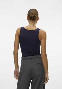 Canotta blu navy con spalline sottili, realizzata in un tessuto liscio, abbinata a pantaloni grigi sartoriali con tasche posteriori e una vestibilità dritta.