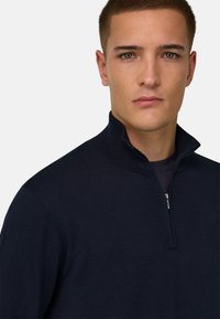 Jeune homme aux cheveux courts portant un pull à fermeture éclair sur le devant de couleur bleu marine foncé, sur un fond clair uni.