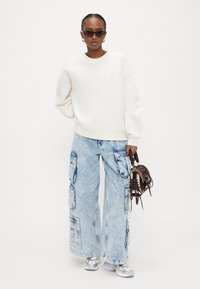 Λευκό φαρδύ φούτερ με ριμπ μανσέτες, συνδυασμένο με ανοιχτόχρωμα, ελαφρά φθαρμένα παντελόνια cargo με φαρδιά μπάτζια και πολλαπλές τσέπες. Μικρή καφέ τσάντα χεριού.