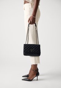 Schwarze, gesteppten Handtasche mit Kettenriemen, ausgestattet mit einem dreieckigen Logo. Getragen zu hellen Jeans und schwarzen Stiletto-Absätzen.