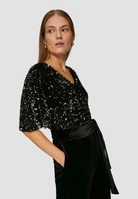 Schwarzes Paillettenoberteil mit V-Ausschnitt und kurzen Ärmeln, kombiniert mit schwarzen Samt-Hosen mit hoher Taille, die einen gebundenen Bund und Taschen aufweisen.