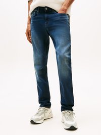 Los jeans de mezclilla azul oscuro presentan un corte recto, un desvanecimiento sutil y un diseño tradicional de cinco bolsillos, combinados con zapatillas deportivas grises.