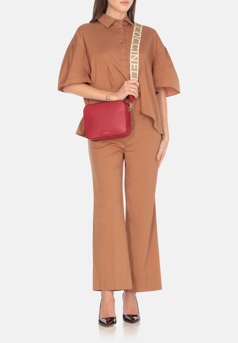 Borsa a tracolla rossa in pelle con finitura testurizzata e una tracolla beige con il nome del brand in grassetto. Forma quadrata e hardware in metallo.