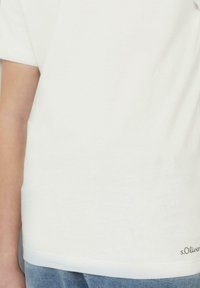 Eine Person trägt ein schlichtes weißes T-Shirt mit dem "s.Oliver"-Logo am Saum, kombiniert mit blauen Jeans, sichtbar von der Taille bis zur Mitte des Arms.