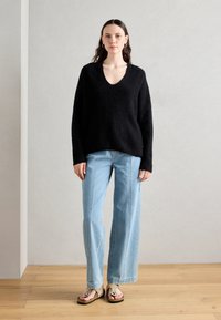 Zwarte gebreide trui met V-hals, losse fit en een gemêleerde afwerking, gecombineerd met lichtblauwe wijde jeans en beige sandalen.