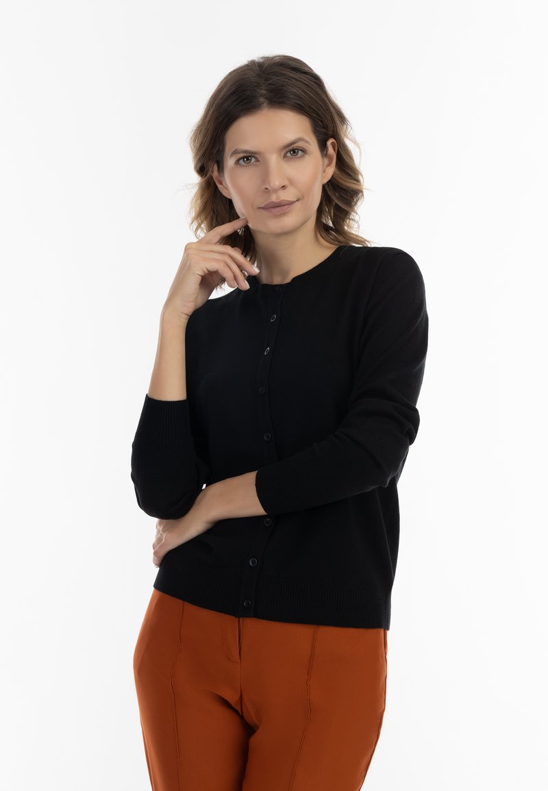 Cardigan nero con maniche lunghe, scollatura rotonda e chiusura con bottoni. Indossato con pantaloni arancioni, evidenziando una combinazione di colori a contrasto.