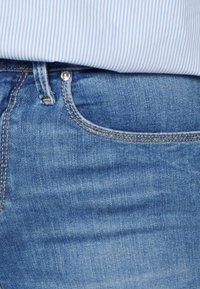Primer plano del bolsillo de unos jeans de mezclilla azul con costura naranja, combinado con una camisa a rayas azul y blanca metida por dentro.