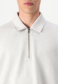 Light grey sweatshirt with a zippered collar, featuring a soft texture and simple design, focusing on the zipper detail at the neck.