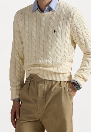 Mann trägt cremefarbenen, kabelgestrickten Pullover über einem blauem Hemd mit Kragen, khakifarbene, hochtaillierte Hose und eine Armbanduhr mit braunem Lederarmband.