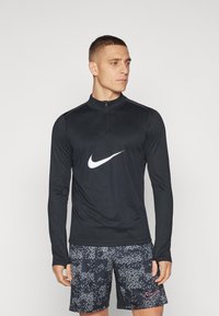 Uomo in cima a maniche lunghe nera con zip con il logo Nike bianco e pantaloni corti neri con motivo camo digitale e logo Nike rosa.
