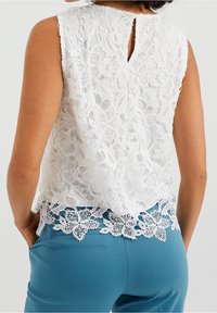 Valkoinen pitsipaita, jossa on kukkakuosi, hihaton malli, selässä avaus, jossa on avainaukko ja pitsireunus helman kohdalla.