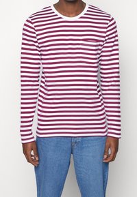 Mann, der ein langärmliges weißes und kastanienbraunes gestreiftes Hemd mit einer Brusttasche und hellblauen Jeans trägt, vor einem schlichten Hintergrund steht.