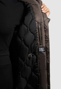 Brun pufjakke med en kviltet sort foer, lynlåslomme og et logomærke indeni. Stoffet ser glat og skinnende ud.