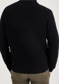 Pull noir en maille avec un col rond, tissu texturé et ourlet côtelé, associé à un pantalon de couleur claire.