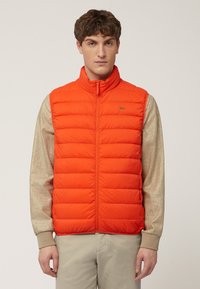 Chaleco acolchado naranja con cuello alto, cremallera frontal y pequeño logo. Combinado con una camisa de manga larga en color beige y pantalones de tono claro.