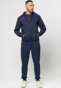 Tumesinine jagatav kapuutsiga pluus, millel on taskud, koos sobiva trenniüksusega. Valmistatud pehmest materjalist, millel on kerged logosümbolid.