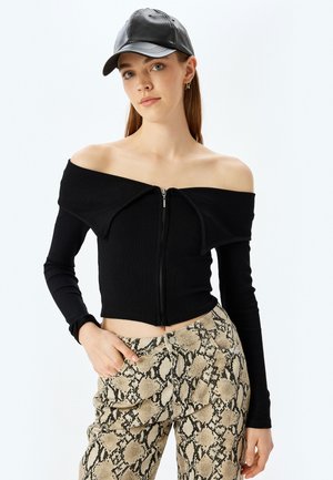 Czarny prążkowany top typu off-shoulder z zamkiem z przodu, połączony z beżowymi spodniami w wężowy nadruk. Ubranie charakteryzuje się długimi rękawami i dopasowanym krojem.