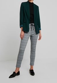 Vihreä bleiseri, jossa on lovetut läpäiset, yhdistettynä harmaan ruudullisiin housuihin ja mustiin loafer-kenkiin. Pehmeä kangas, slim fit ja käännetyt hihat bleiserissä.