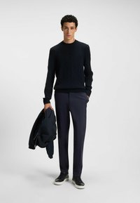 Marineblaue Pullover mit Kabelstrick, kombiniert mit schmal geschnittenen marineblauen Hosen und hält eine marineblaue Jacke. Die Schuhe weisen schwarz-weiße Akzente auf.